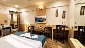 A hotel room with a bed, a desk, two chairs, a small table, a painting on the wall, and a kitchenette. - Hotel Shree Residency (Agra)
