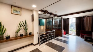 Eine Hotelhalle mit einer Rezeption, Zimmerpflanzen, einer Treppe und Blick auf die Stadt durch große Fenster.