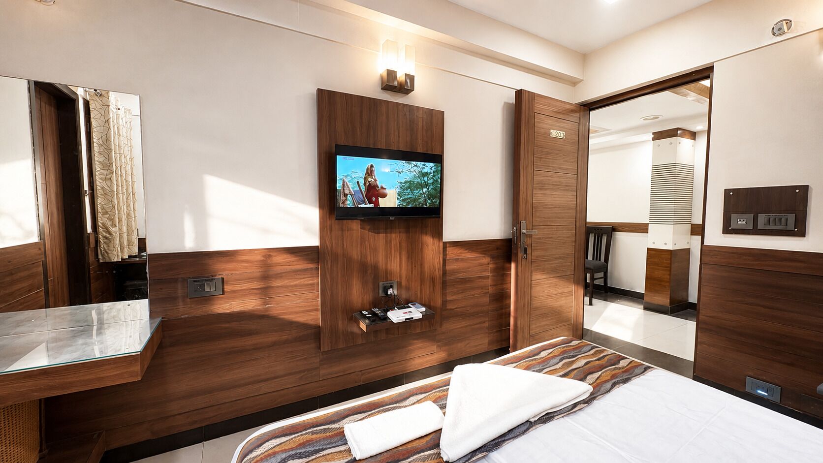 Una habitación de hotel moderna con una cama, un televisor montado en un panel de madera, una tocador con espejo y un armario.