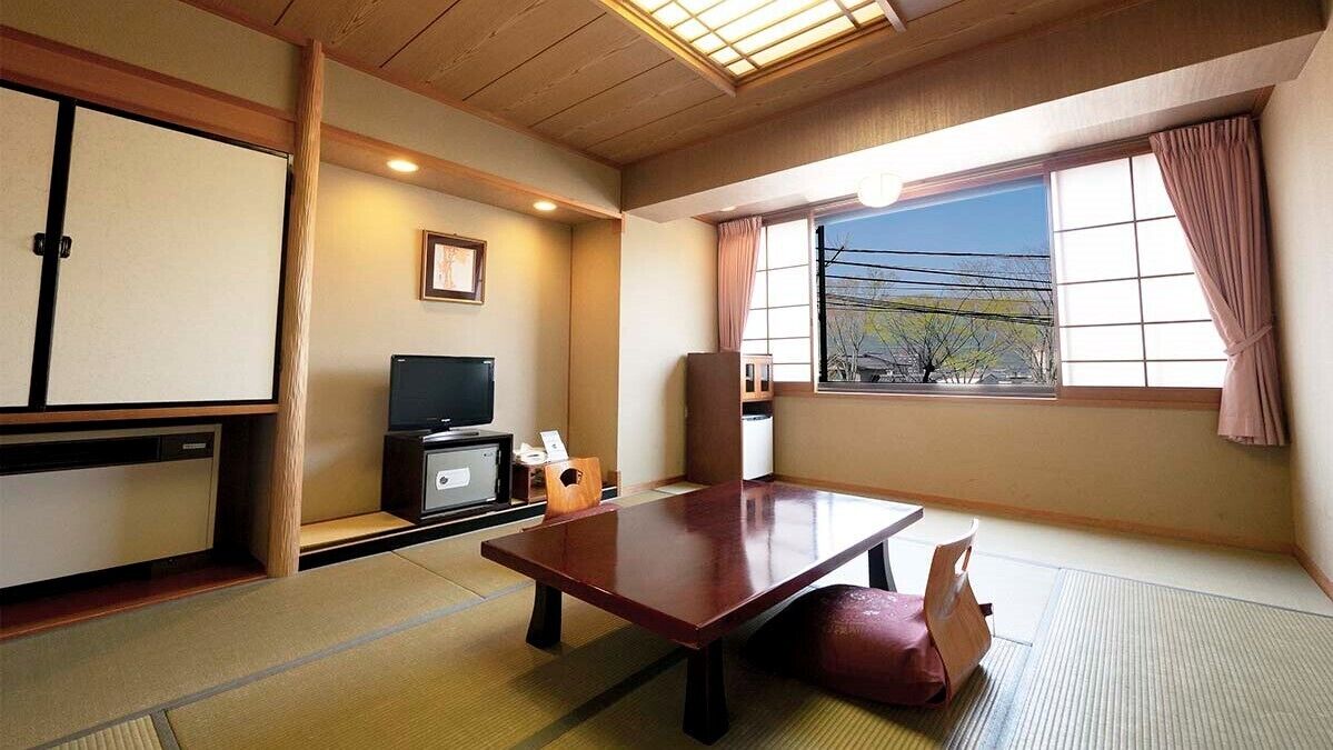 Ein traditioneller japanischer Raum mit Tatami-Böden, einem niedrigen Tisch, einem Fernseher und einem Fenster mit Blick nach draußen.