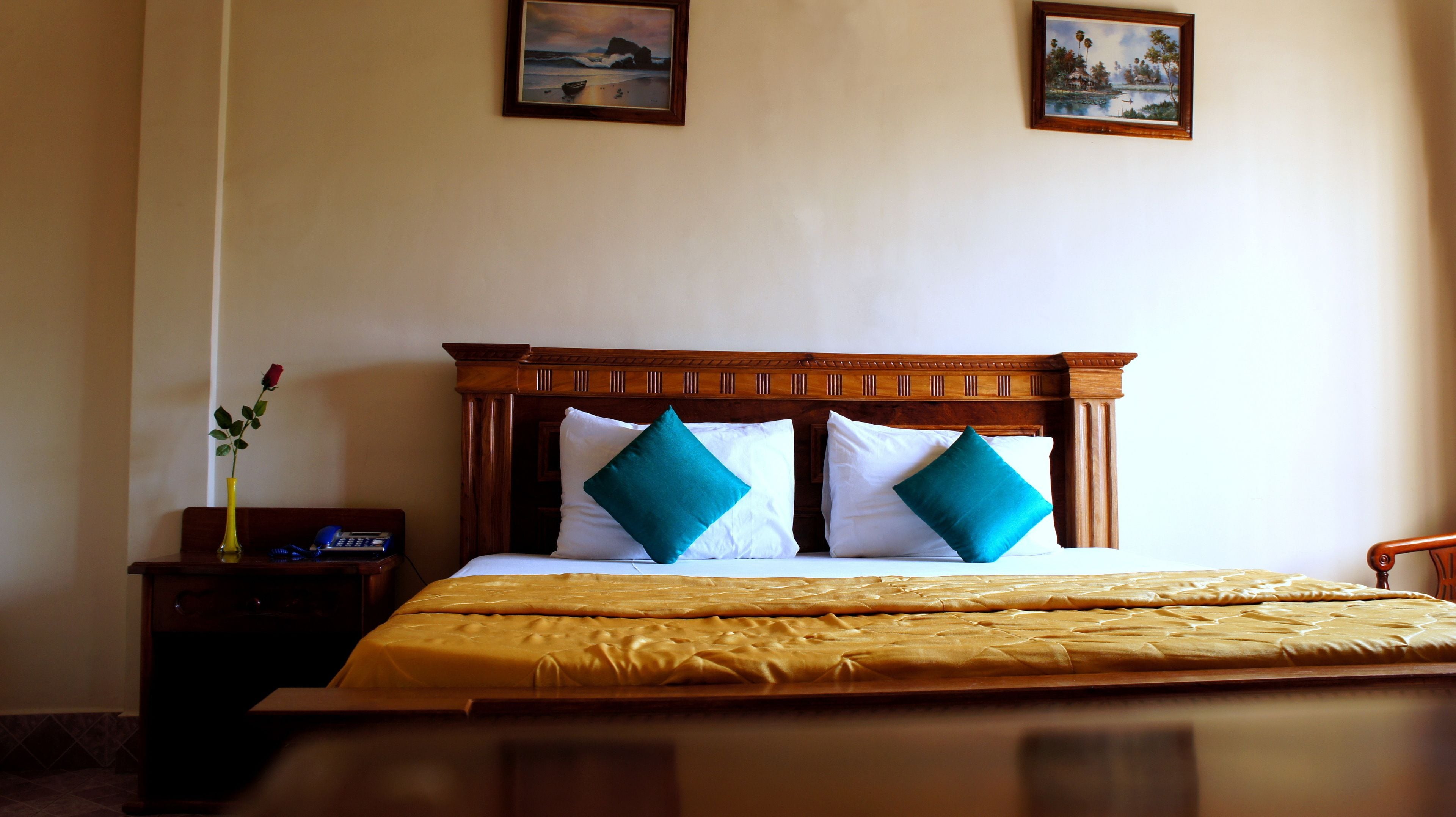 A hotel room with a wooden headboard, two white pillows, a teal pillow, a yellow vase with a red rose, and a small table with a phone and a lamp.