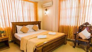 A neatly made bed with a wooden headboard, two pillows, and a beige bedspread. There is a wooden nightstand with a lamp and a small plant beside the bed. A wooden chair with a cushion is positioned next to the bed. - Star Hotel (Battambang)
