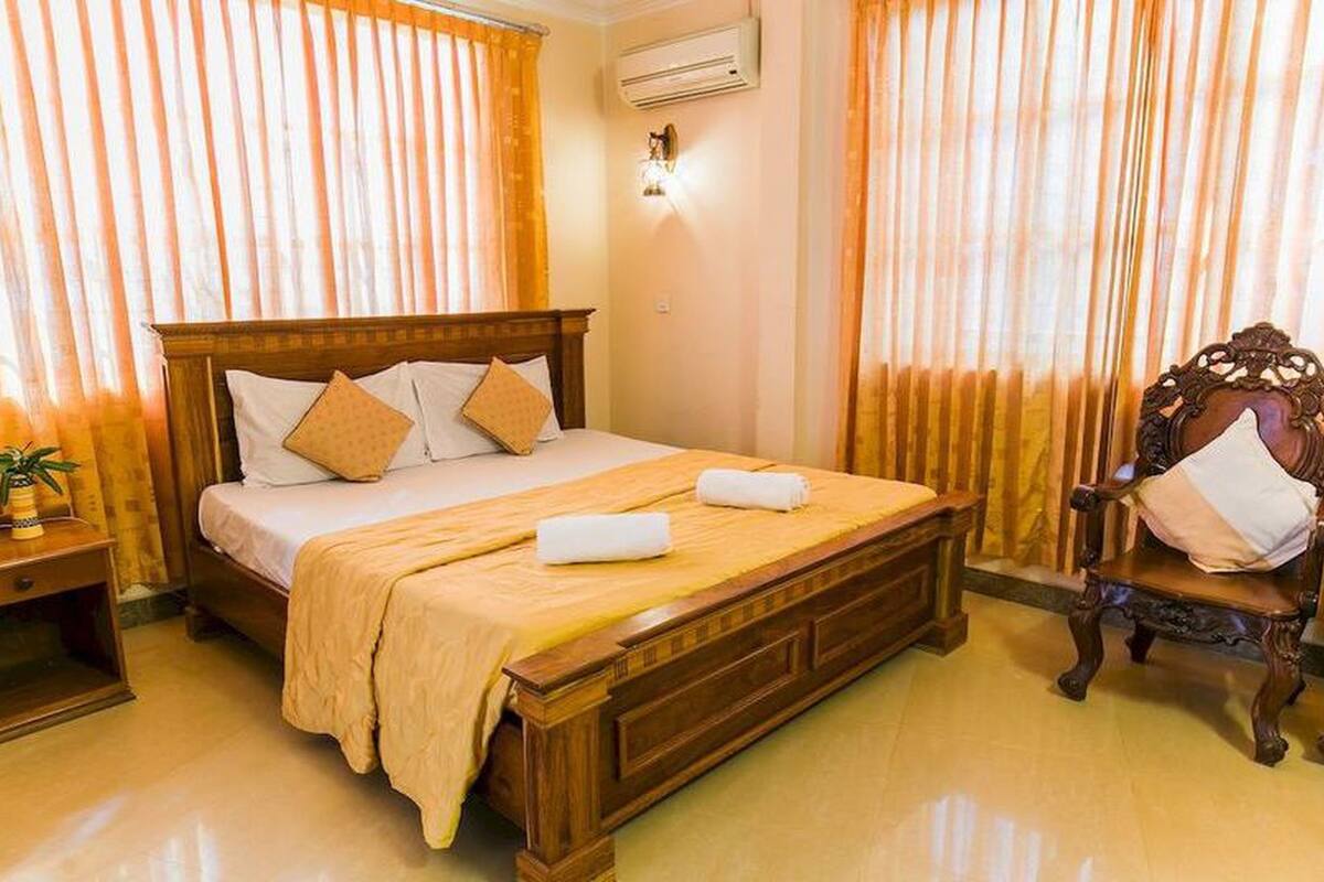 A neatly made bed with a wooden headboard, two pillows, and a beige bedspread. There is a wooden nightstand with a lamp and a small plant beside the bed. A wooden chair with a cushion is positioned next to the bed.