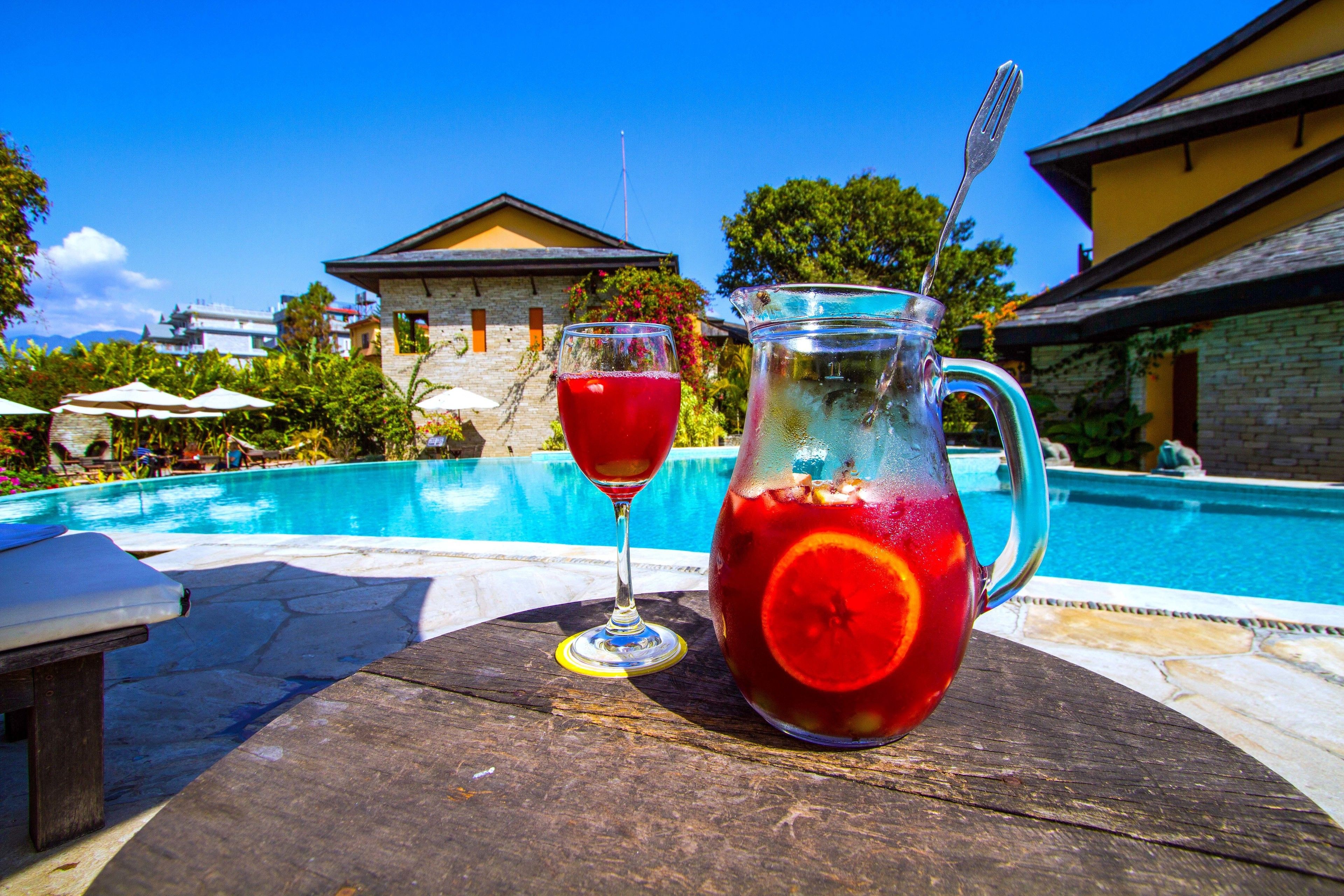 Una mesa junto a la piscina con una jarra y un vaso de bebida roja, una piscina y un edificio al fondo.