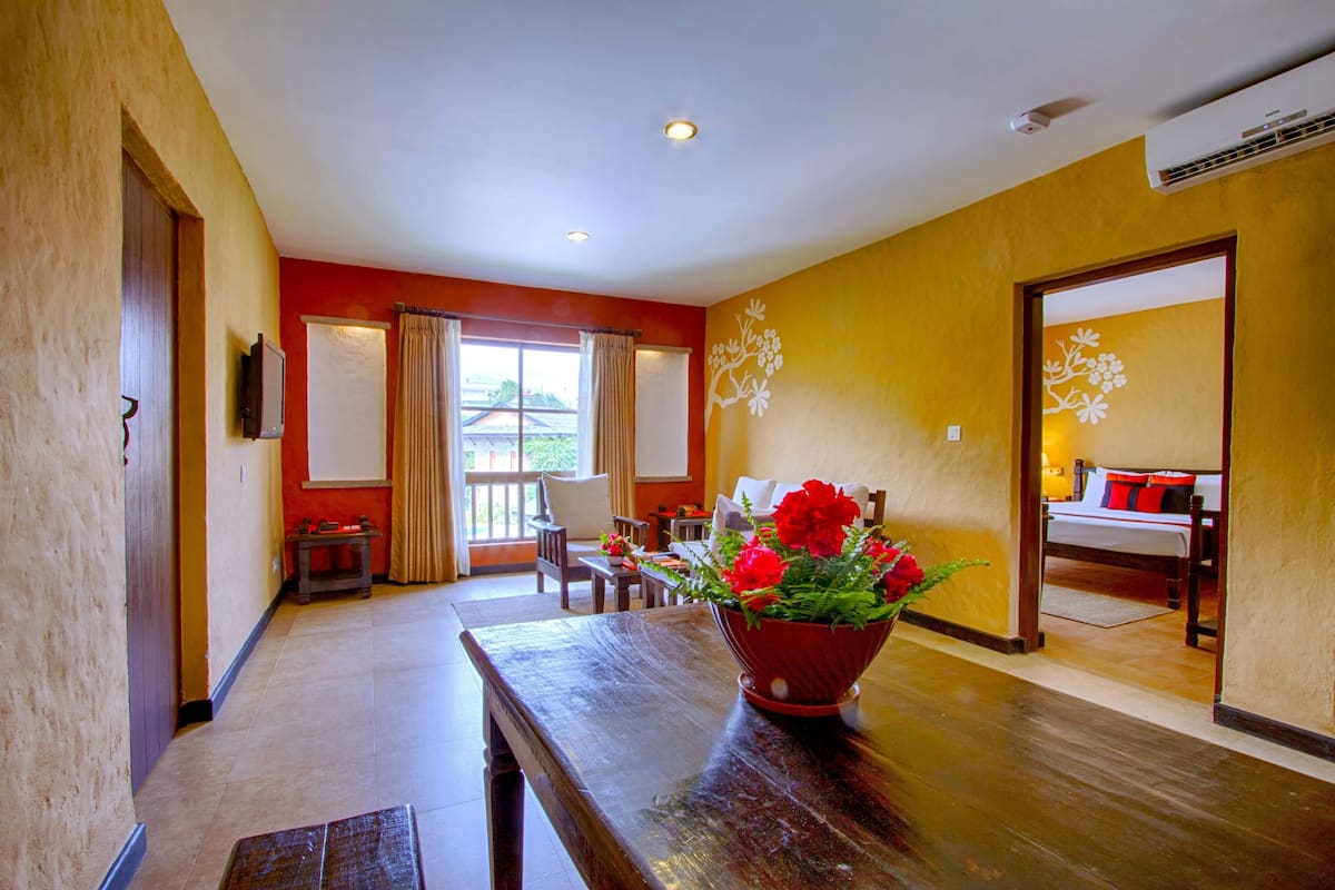 A hotel room with a wooden table, a red flower vase, a bed with red and white bedding, and a window with curtains.