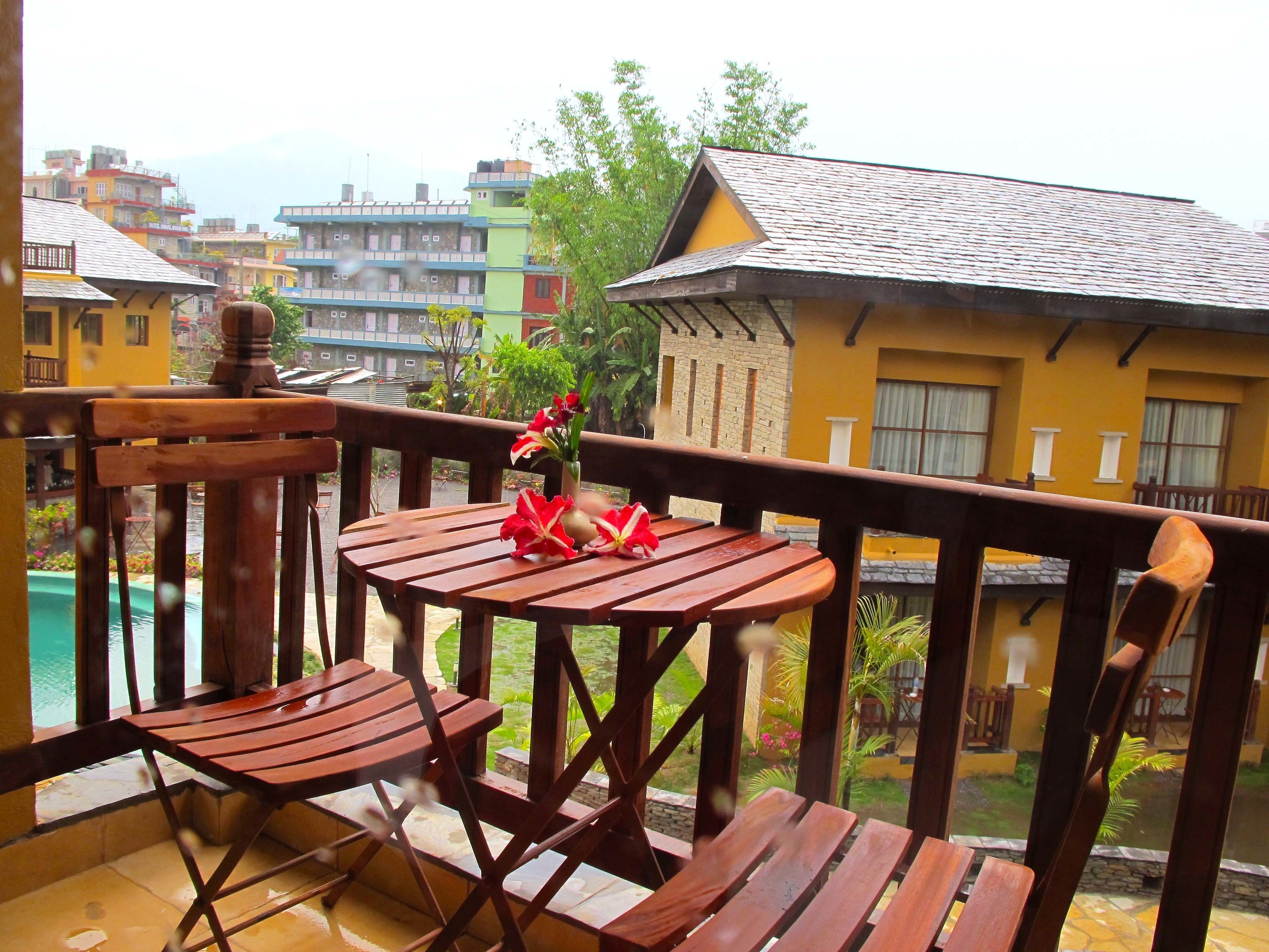 A wooden balcony with a table and chairs, overlooking a pool and colorful buildings.