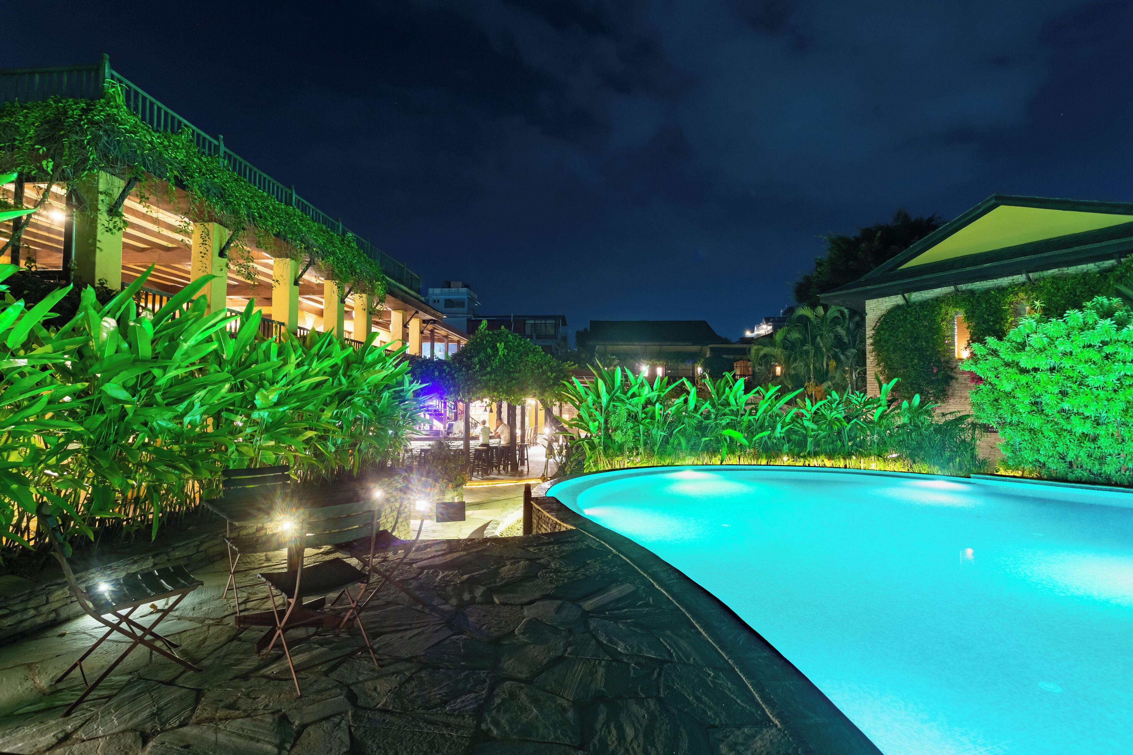 Vue nocturne d’un hôtel avec une piscine, un chemin de pierre et une végétation luxuriante.