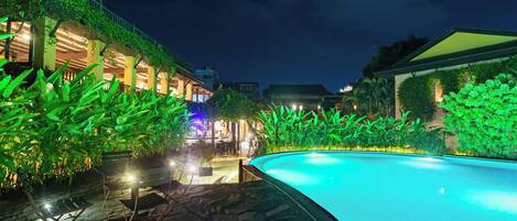 Vue nocturne d’un hôtel avec une piscine, un chemin de pierre et une végétation luxuriante.