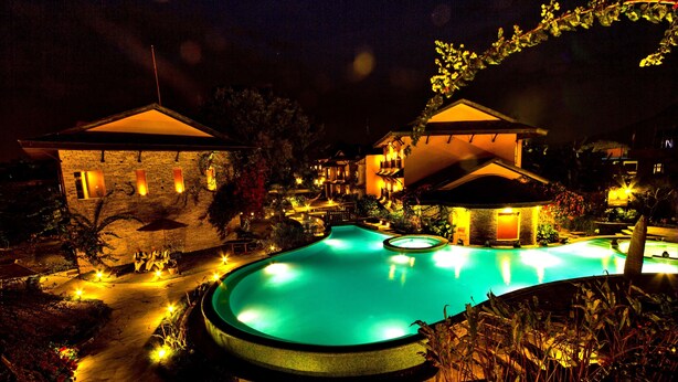 Una piscina rodeada de edificios con iluminación cálida, un camino de piedra y jardines ajardinados.