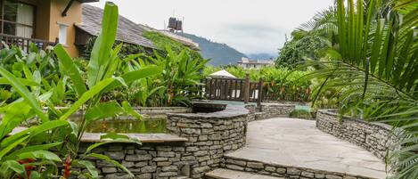 A stone pathway leading to a pond, surrounded by lush greenery and a building with a balcony.