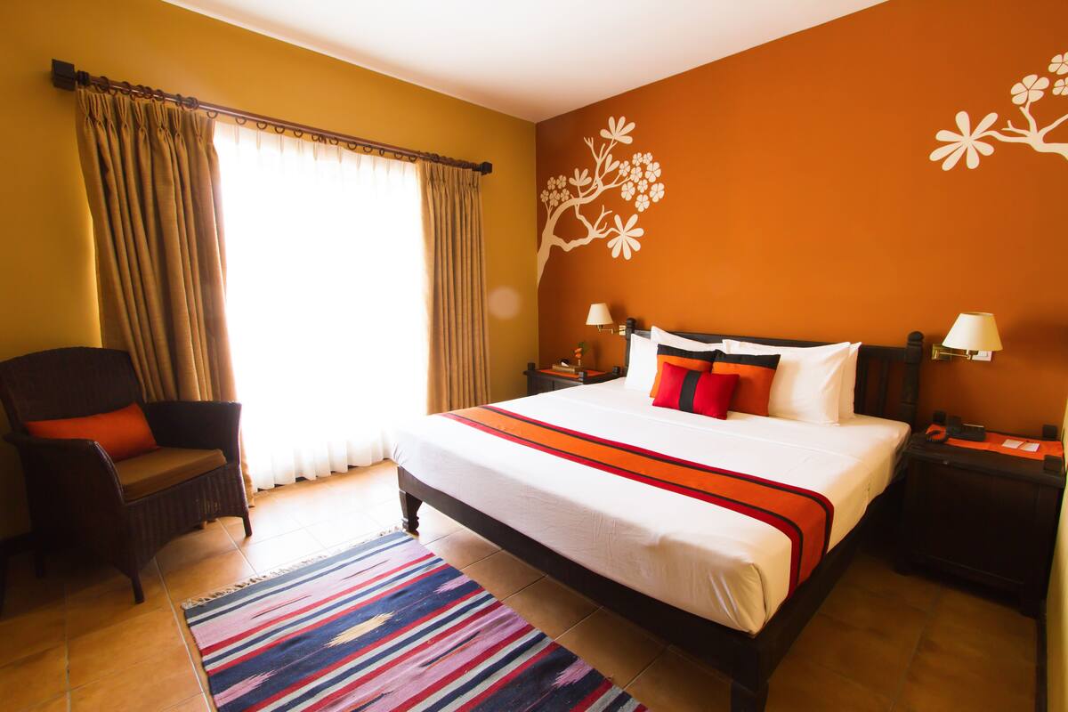 A hotel room with a bed, a chair, a nightstand, a lamp, and a window with curtains.