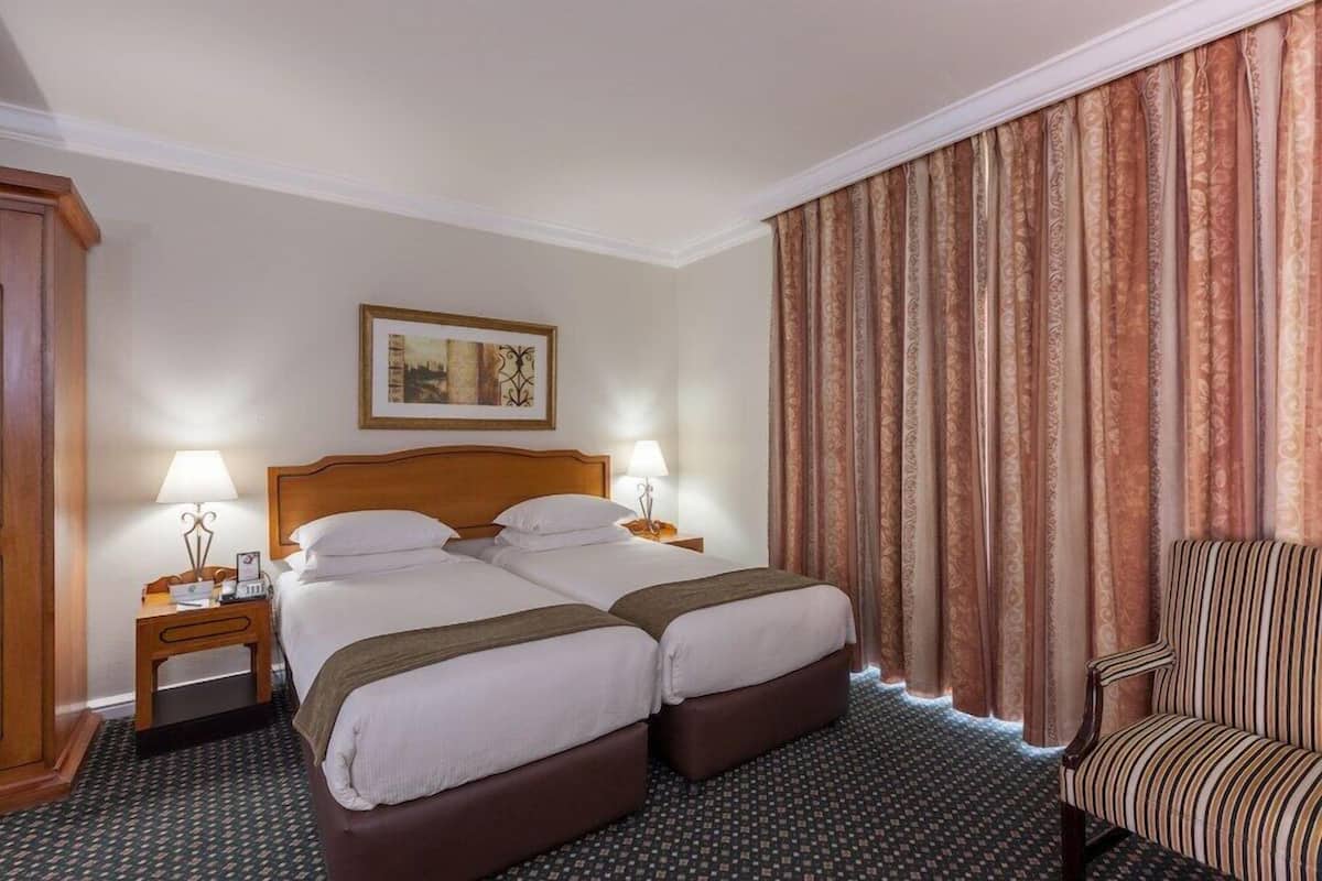 Ein Hotelzimmer mit zwei Betten, einem Sessel, einem Nachttisch mit Lampe und einem gerahmten Bild an der Wand.