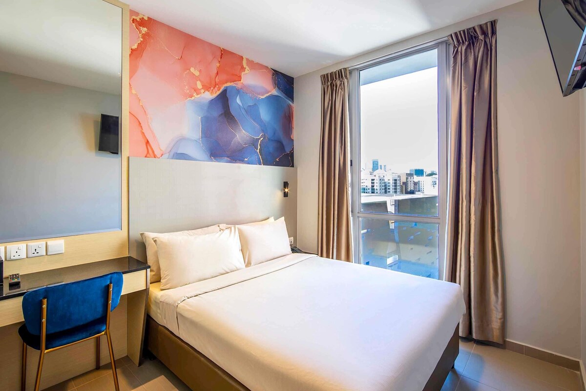 Habitación de hotel con cama, escritorio, silla, ventana grande con cortinas y vista a la ciudad.