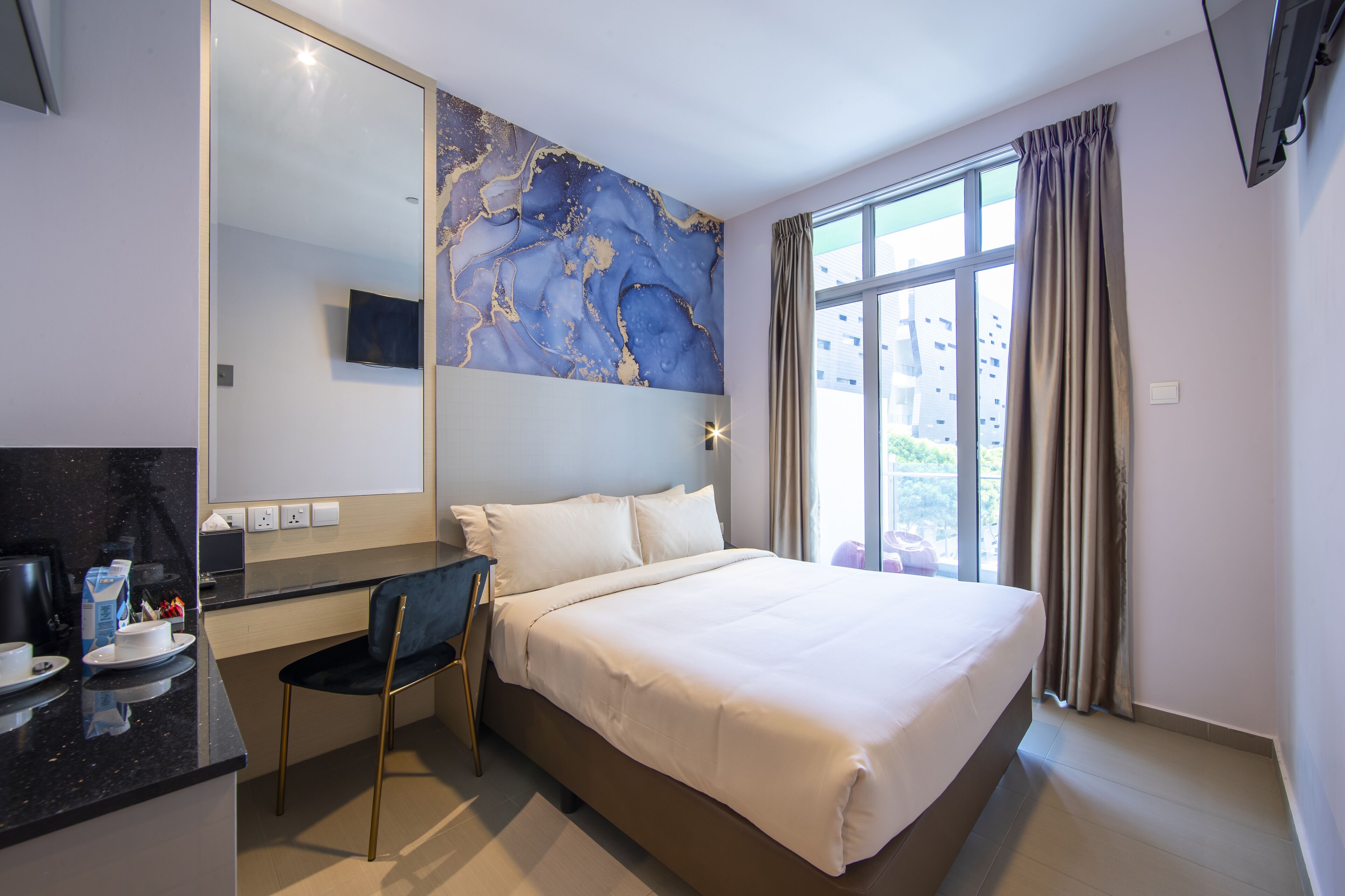 Quarto de hotel com cama, escrivaninha, cadeira, espelho grande e janela com cortinas.