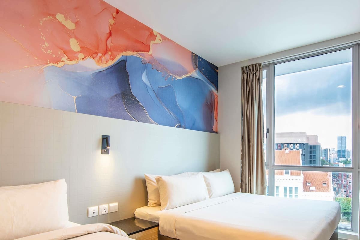 A hotel room with two beds, a large abstract painting on the wall, a window with a city view, and a nightstand with a lamp.