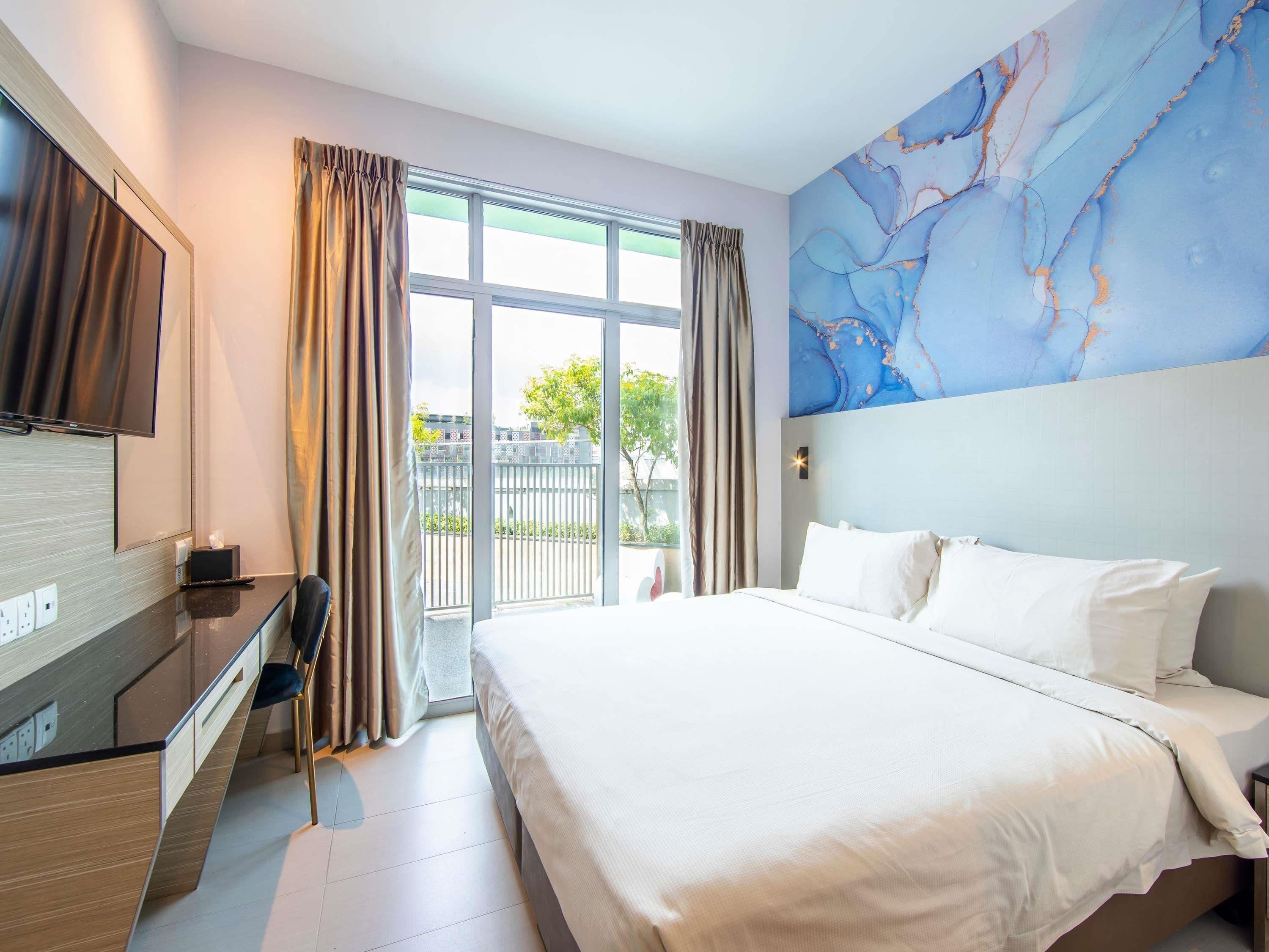 A modern hotel room with a large bed, a flat-screen TV, a desk with a chair, and a view of the outdoors through large windows.