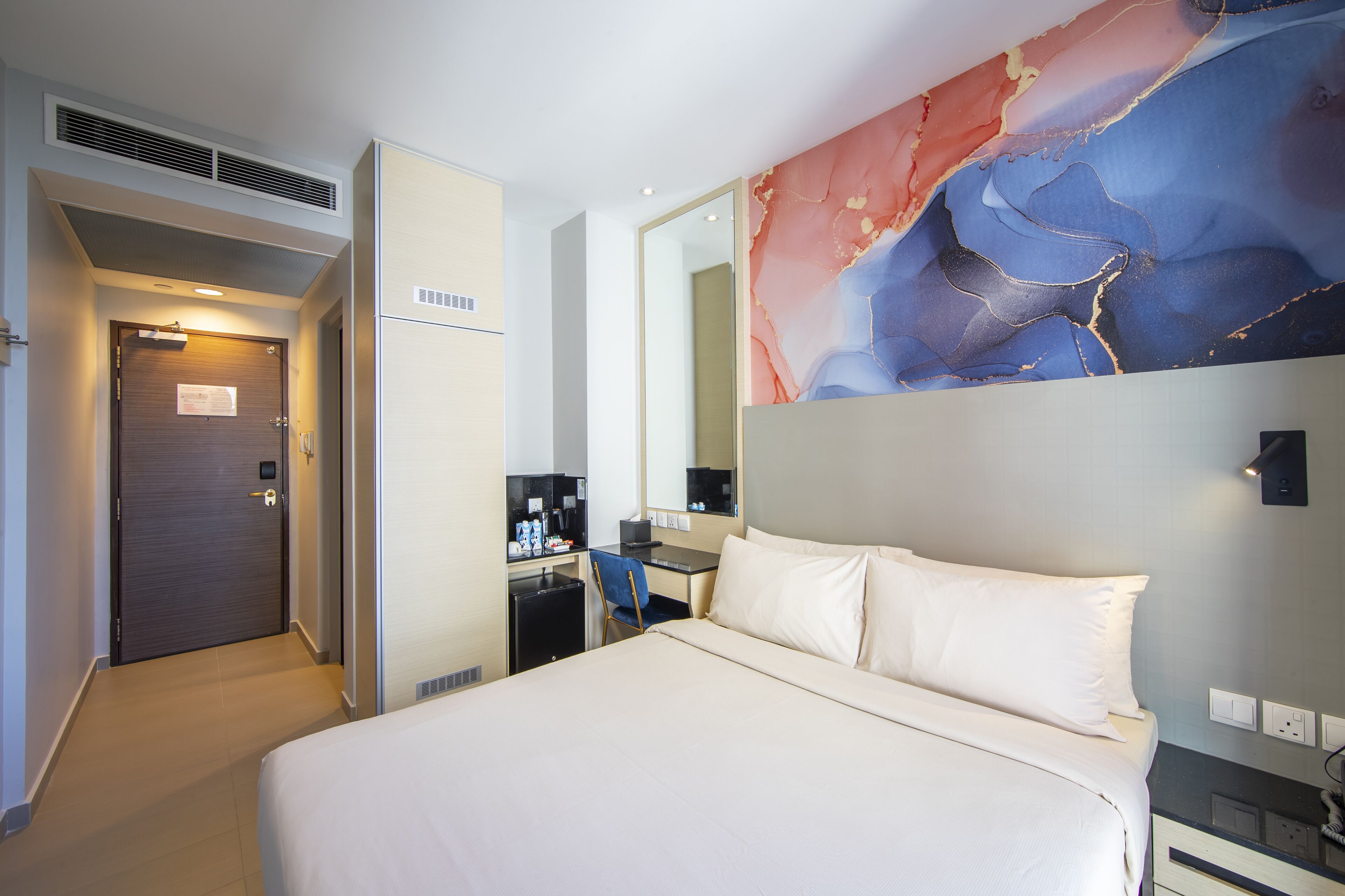 A hotel room with a bed, a desk, a chair, a mirror, and a wall with a colorful abstract design.