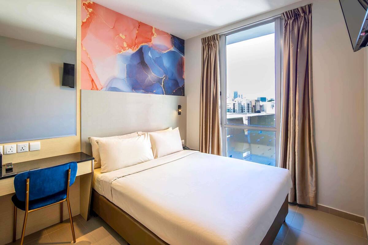 A hotel room with a bed, a desk, a chair, a large window with curtains, and a city view.