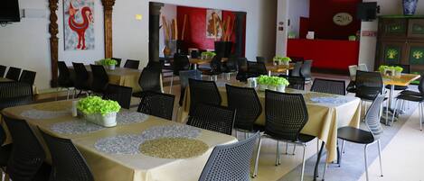 A restaurant with multiple tables and chairs, some with plants. There are decorative elements like a rooster painting and a red wall.
