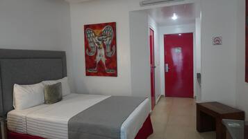 A hotel room with a bed, a painting of an angel, a red door, and a 'No Smoking' sign.