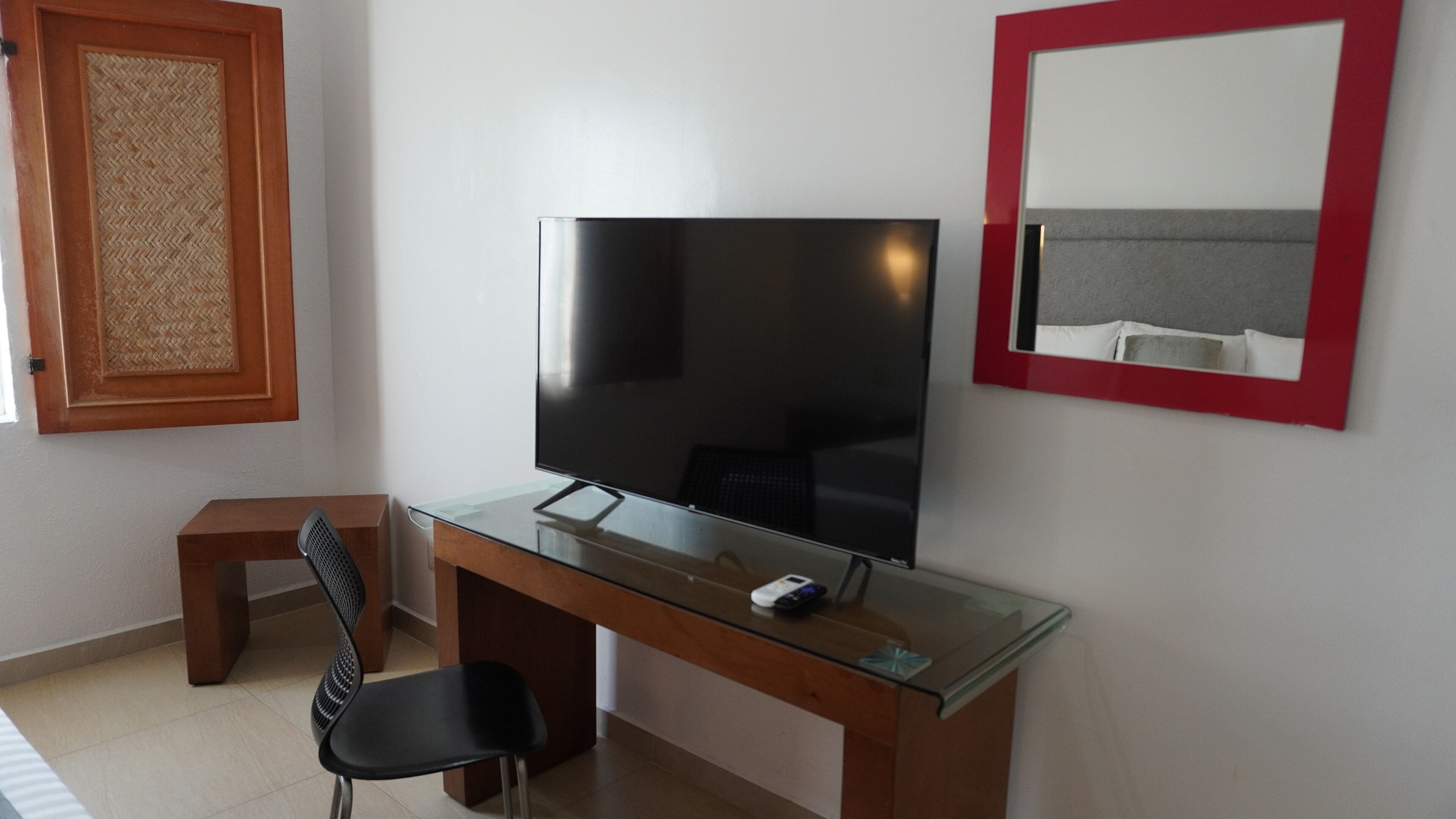 Une chambre moderne avec un téléviseur à écran plat posé sur un bureau en verre, un petit tabouret en bois et un miroir avec un cadre rouge.