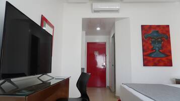 Habitación de hotel con televisión de pantalla plana, un escritorio, una silla, una cama, una puerta roja y un cuadro en la pared.