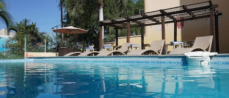 A swimming pool with lounge chairs and umbrellas, surrounded by palm trees and a covered area.