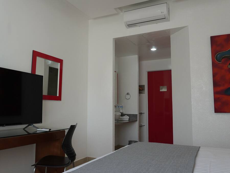 Habitación de hotel con cama, televisor, escritorio con silla, puerta roja y un cuadro en la pared.