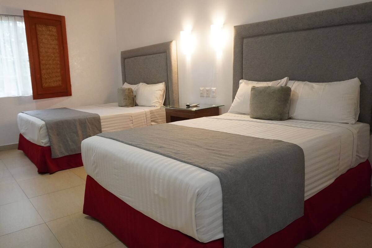 A hotel room with two beds, a large window with curtains, a bedside table with a lamp, and a wall-mounted light fixture.