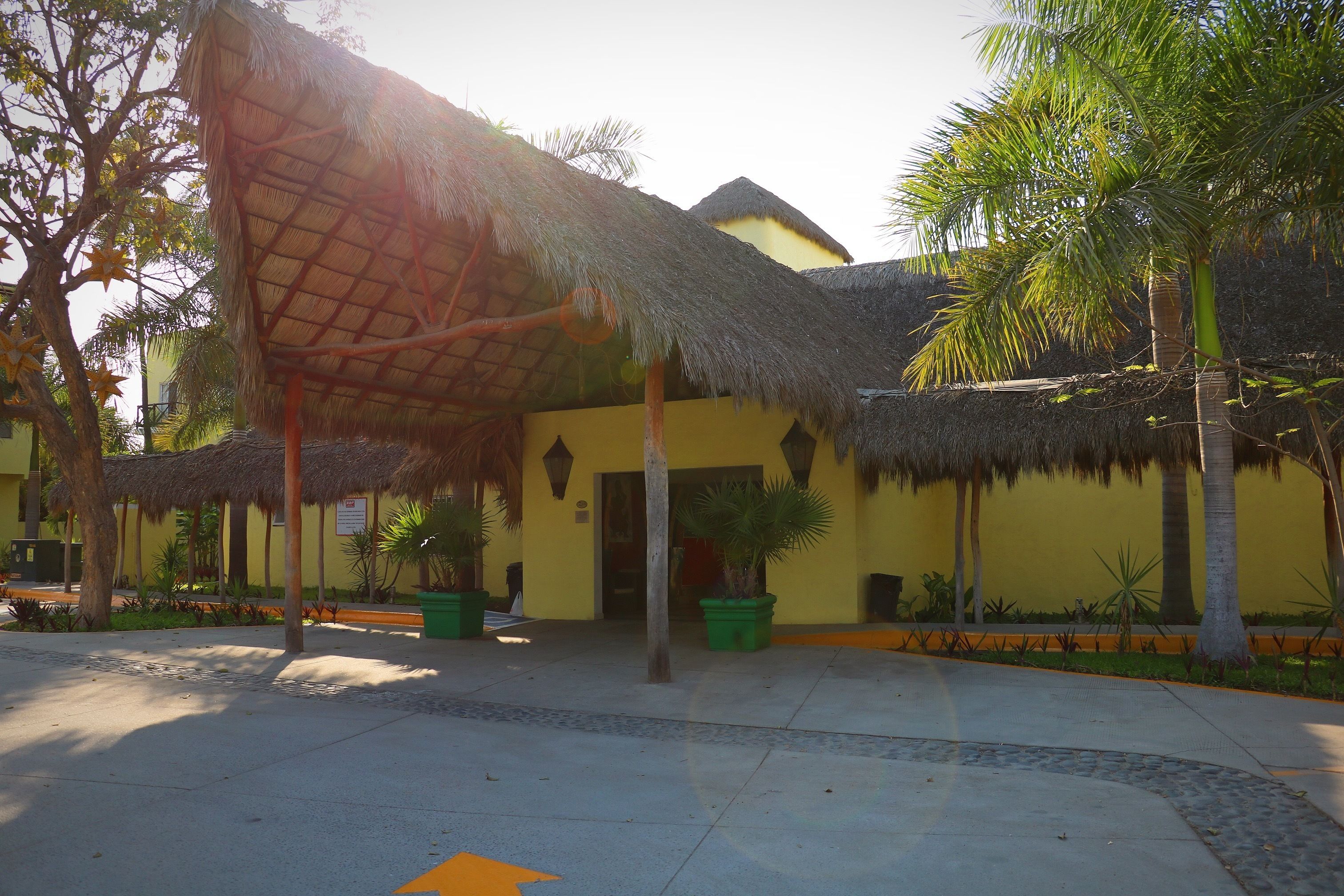 A building with a thatched roof and a yellow facade, surrounded by palm trees and potted plants.