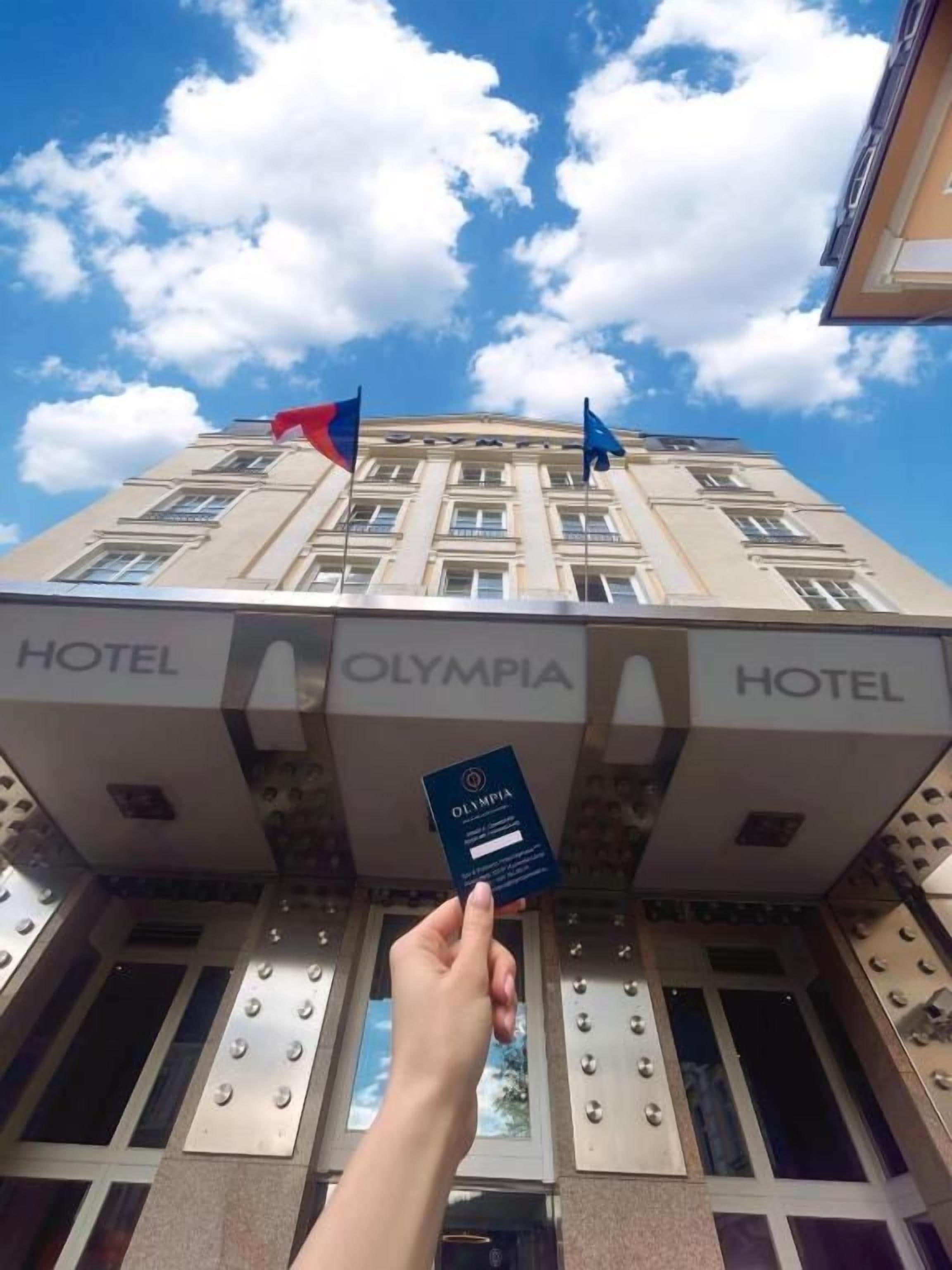 A hand holding a hotel card in front of a building with 'HOTEL' signs and flags.