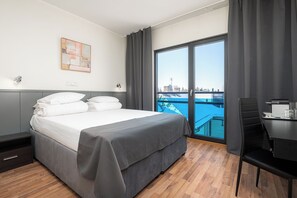 A hotel room with a bed, a desk, a chair, a window with a view of a cityscape, and a painting on the wall.
