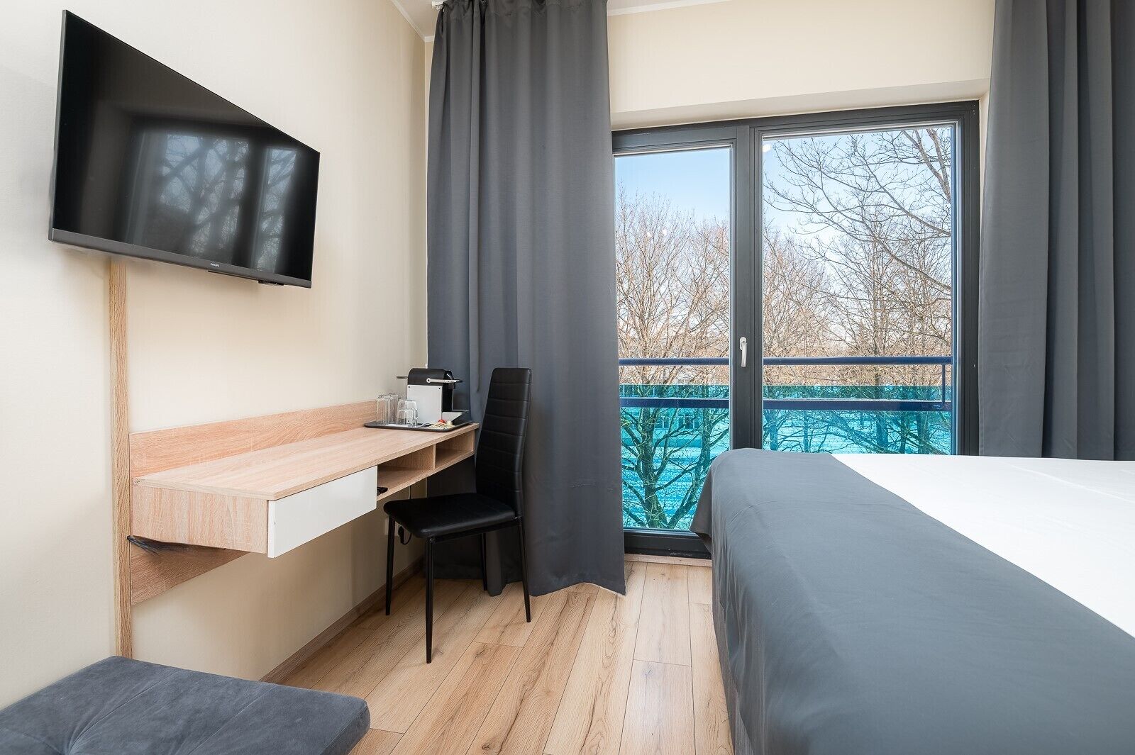 A hotel room with a bed, a desk with a chair, a TV mounted on the wall, and a balcony with a view of trees.