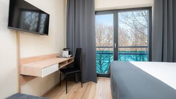 Ein Hotelzimmer mit Bett, Schreibtisch mit Stuhl, Wandfernseher und Balkon mit Blick auf Bäume.
