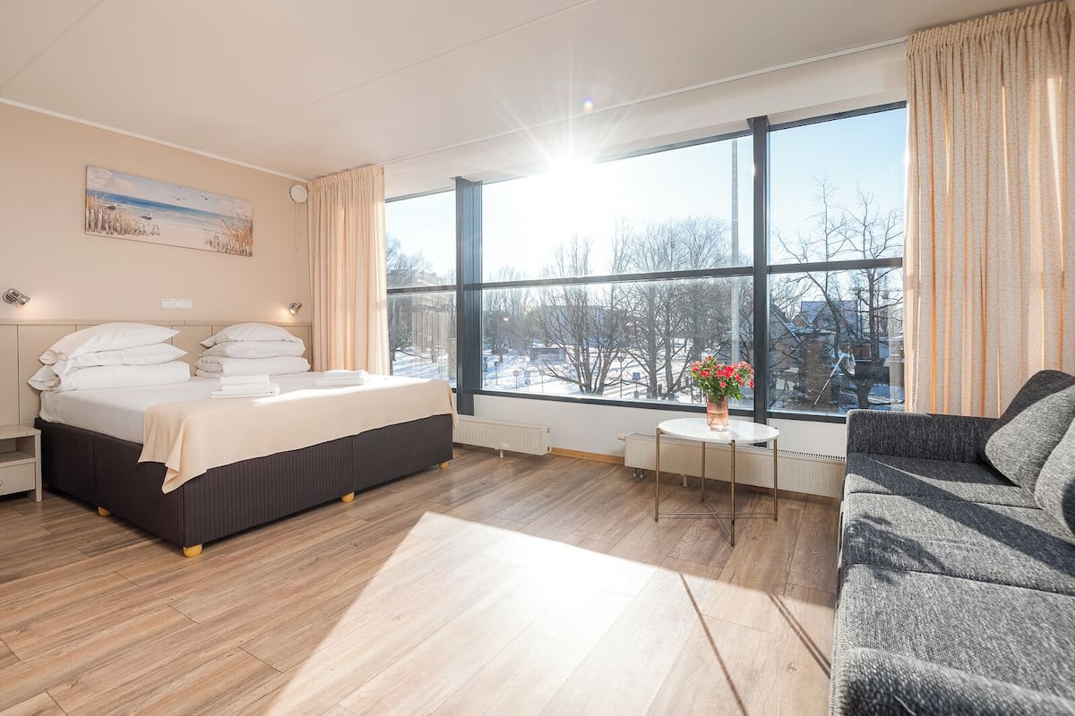 Habitación de hotel con un ventanal grande, una cama, una mesita de noche, una silla y una pequeña mesa con un florero.