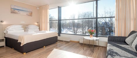 Habitación de hotel con un ventanal grande, una cama, una mesita de noche, una silla y una pequeña mesa con un florero.