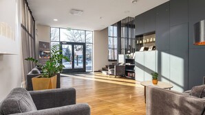 A modern hotel lobby with a wooden floor, grey sofas, a large potted plant, and a bookshelf. - Hotell Tammsaare Full Automated Hotel (Parnu)