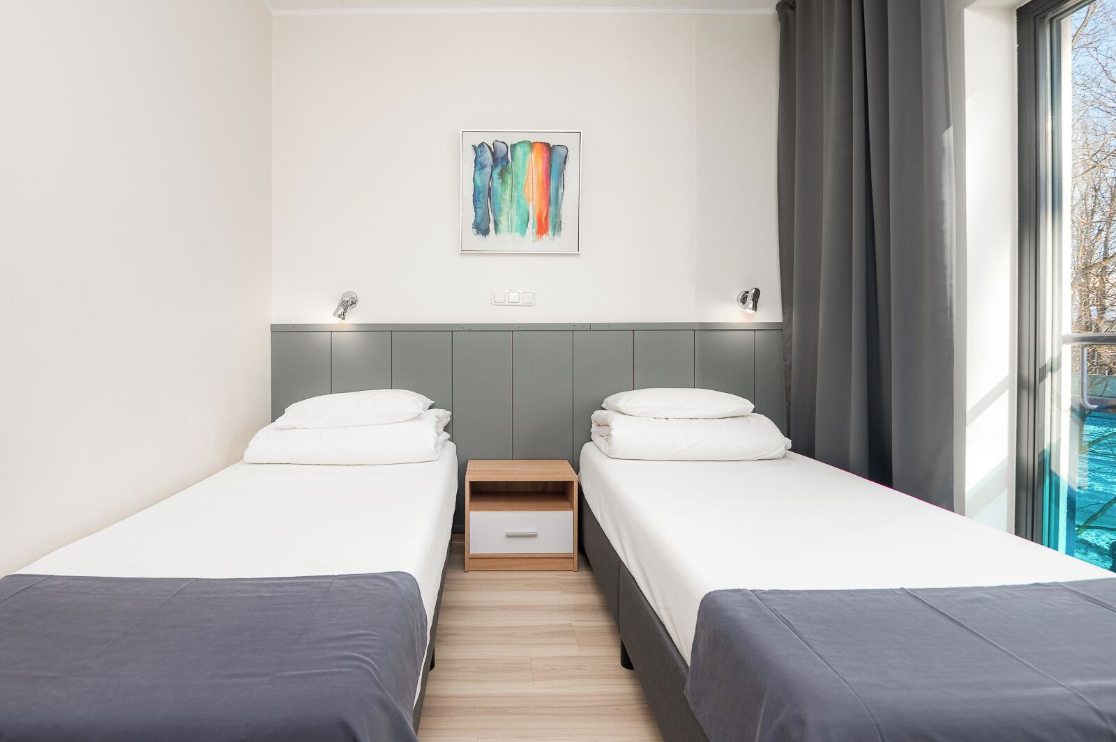 A modern hotel room with two single beds, a wooden nightstand, a small drawer, and a colorful abstract painting on the wall.