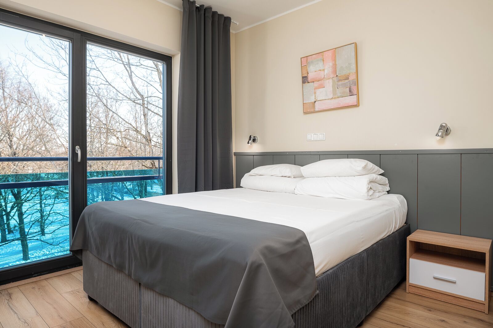 A bedroom with a bed, a nightstand, a window with a view of trees, and a wall-mounted light fixture.