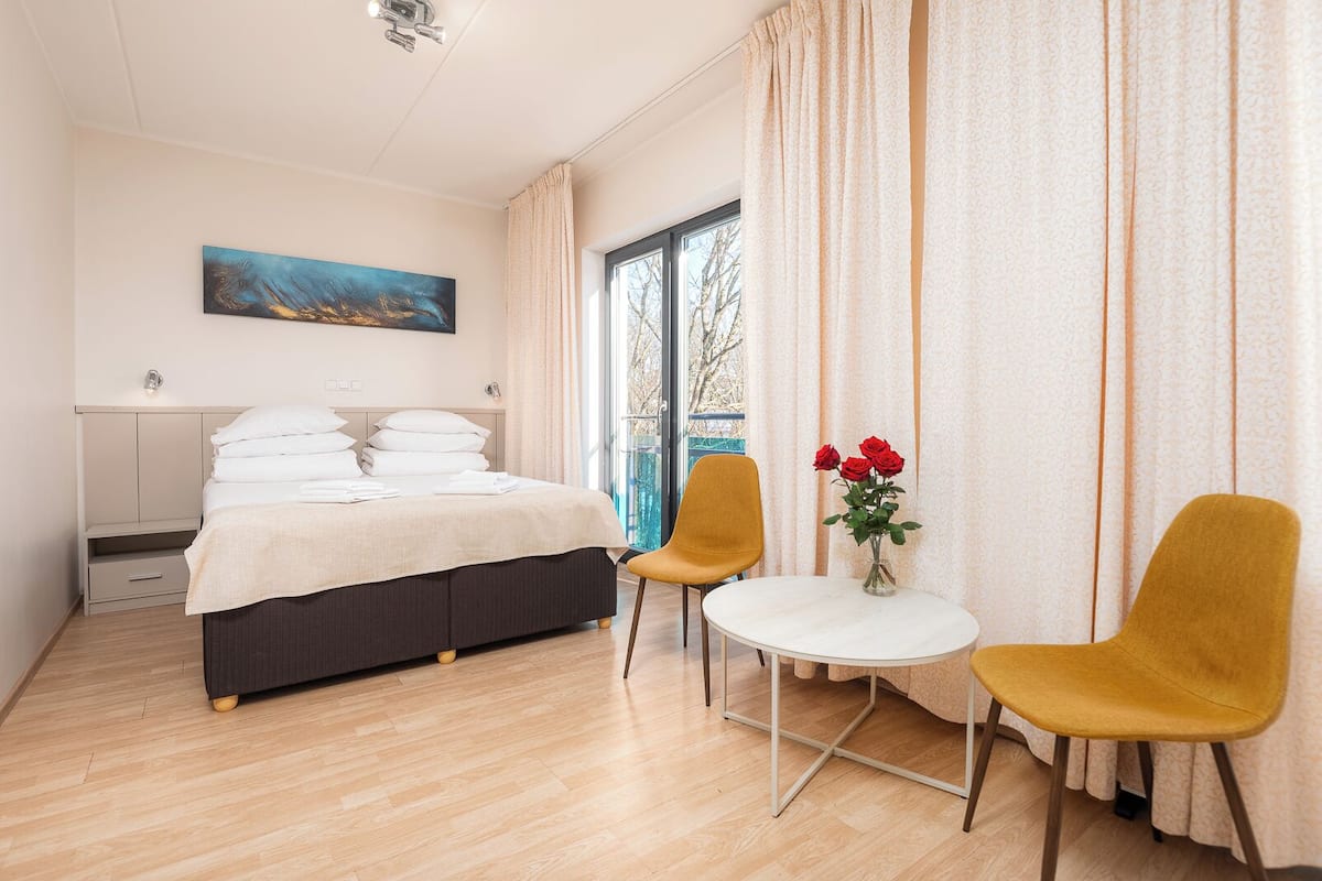 Habitación de hotel con cama, dos sillas, una mesita con flores, un cuadro en la pared y una ventana con cortinas.