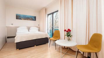 Habitación de hotel con cama, dos sillas, una mesita con flores, un cuadro en la pared y una ventana con cortinas.