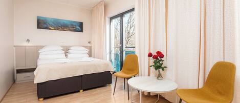 A hotel room with a bed, two chairs, a small table with flowers, a painting on the wall, and a window with curtains.