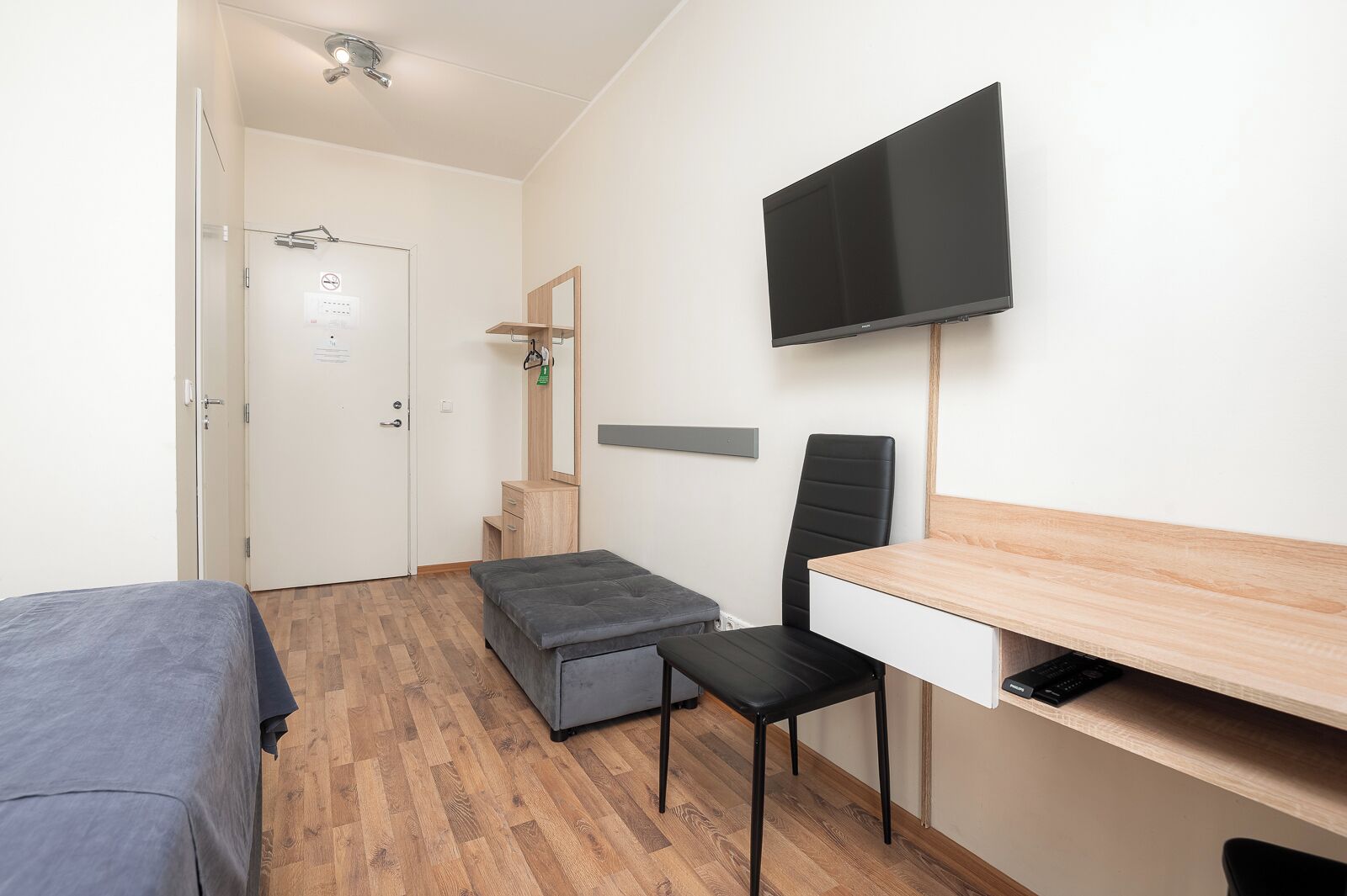 A small, modern room with a bed, a desk, a chair, a television, and a small bench.