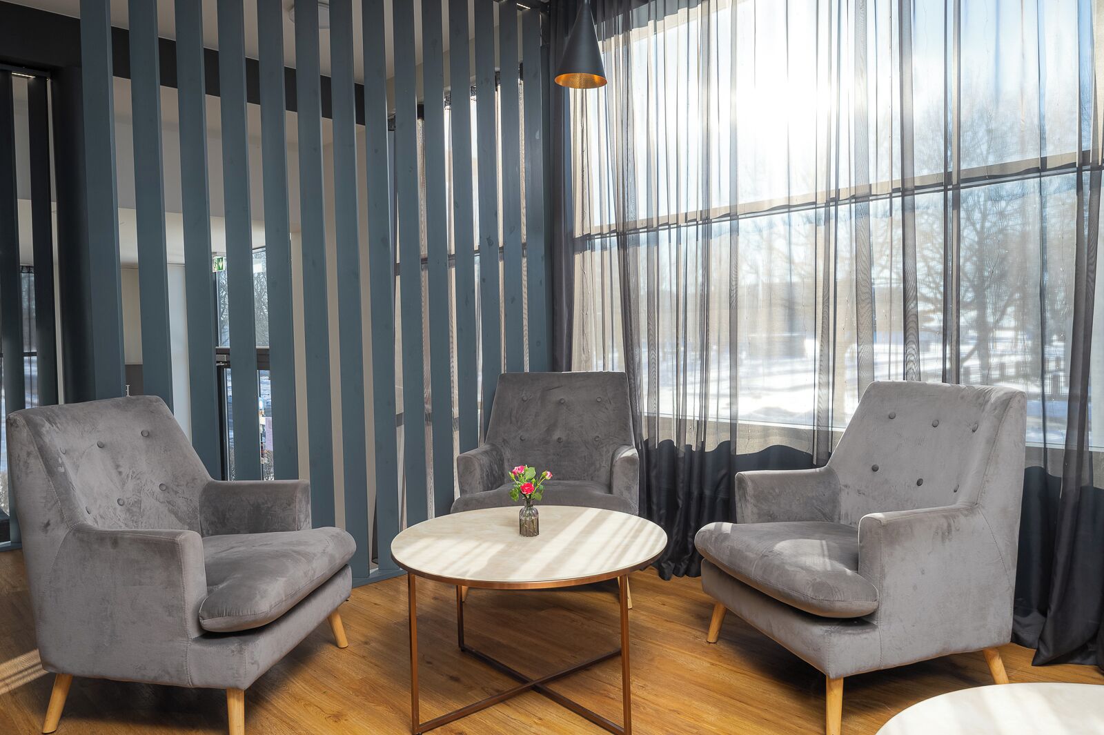 A room with two grey armchairs, a round wooden table with a vase of flowers, and a large window with sheer curtains.