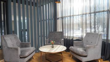 A room with two grey armchairs, a round wooden table with a vase of flowers, and a large window with sheer curtains.