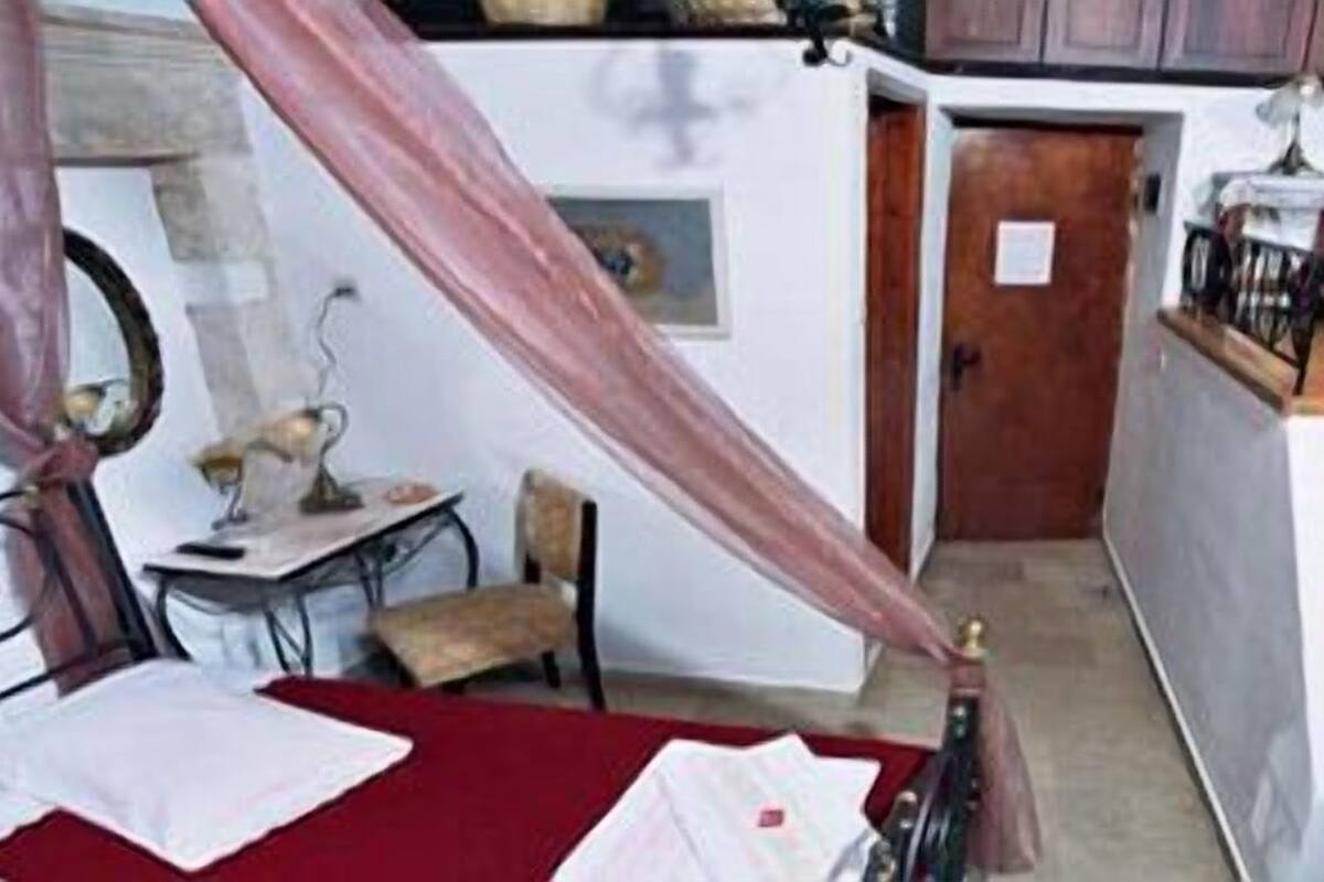 A compact room with a bed, a desk, and a chair. There is a small table with a lamp, a mirror, and a window with curtains. A wooden ladder is visible, leading to a higher level.