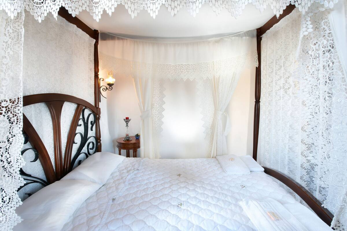 A bedroom with a four-poster bed, lace curtains, and a small bedside table with a lamp.