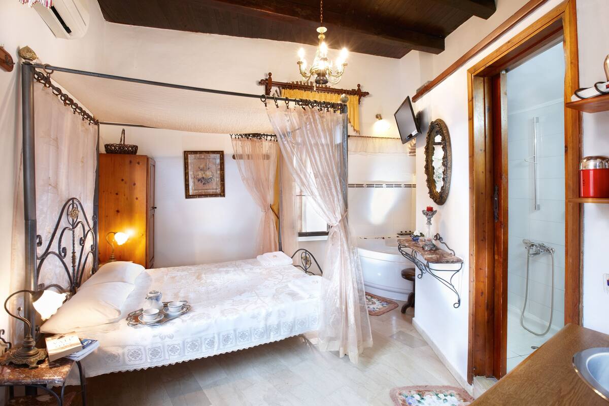 A bedroom with a four-poster bed, a bedside table with a lamp, a small table with a candle, and a window with curtains.