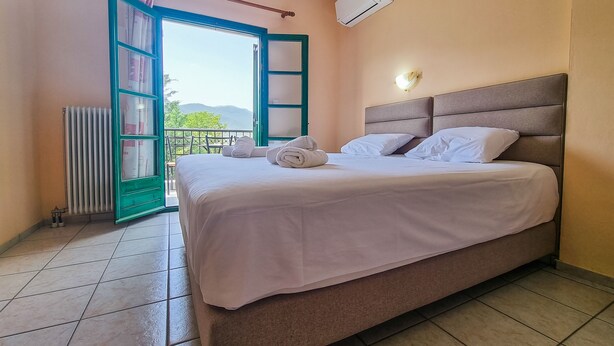 A neatly made bed with white linens and pillows, a radiator, a window with a view of greenery, and an air conditioning unit on the wall. - Hotel Spiridoula (Konitsa)