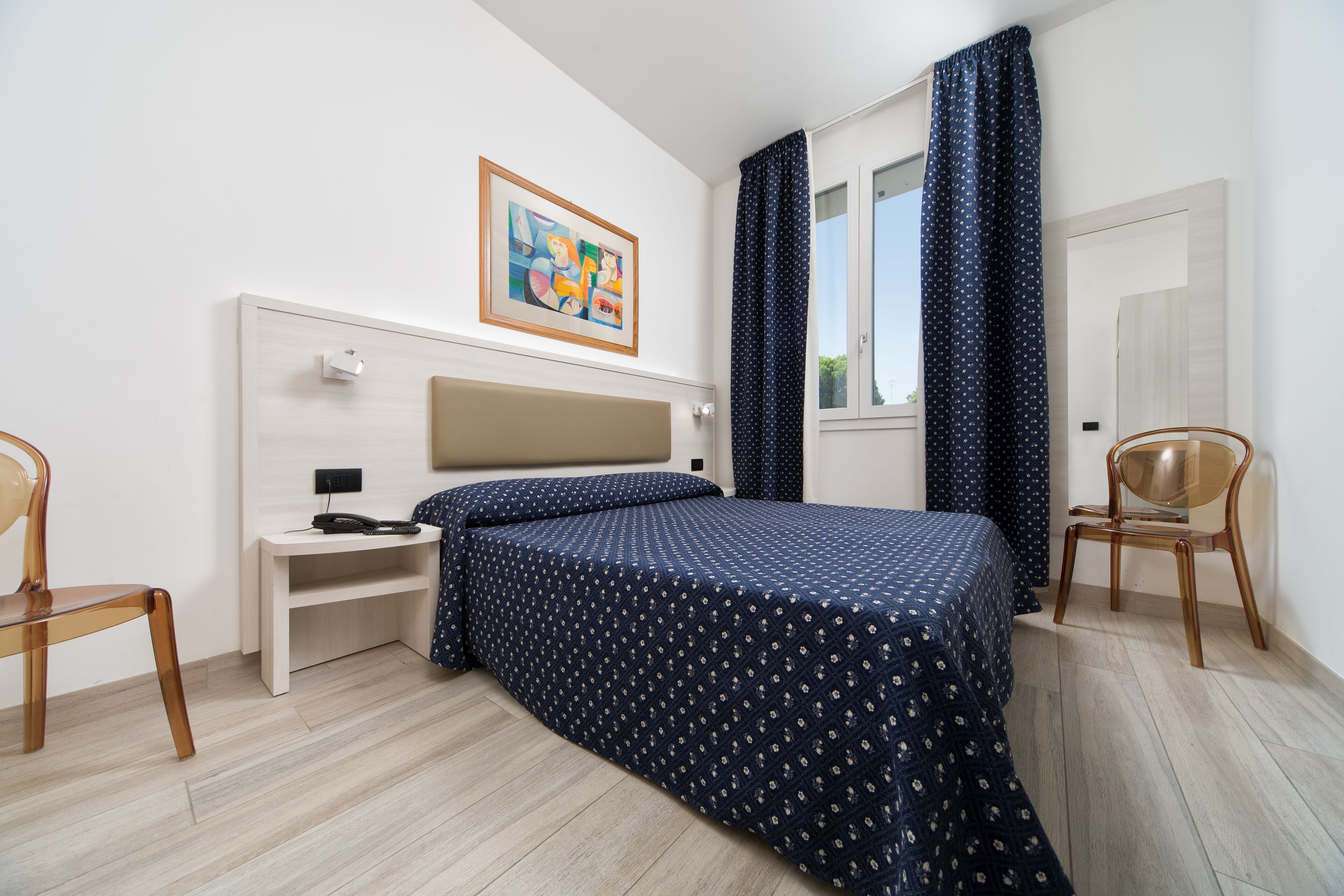 A bedroom with a bed, a nightstand, a chair, a window with curtains, and a painting on the wall.